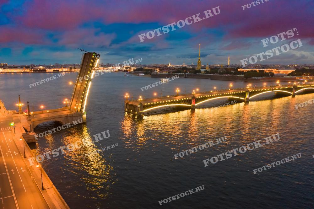 На фотографии изображён разведённый Троицкий мост в Санкт-Петербурге. Игра света от фонарей моста на фоне вечернего неба и тихой поверхности Невы создает завораживающий городской пейзаж. Эта фотография прекрасно подходит для статей о ночной жизни города, о его инженерных достижениях и архитектурных ориентирах.